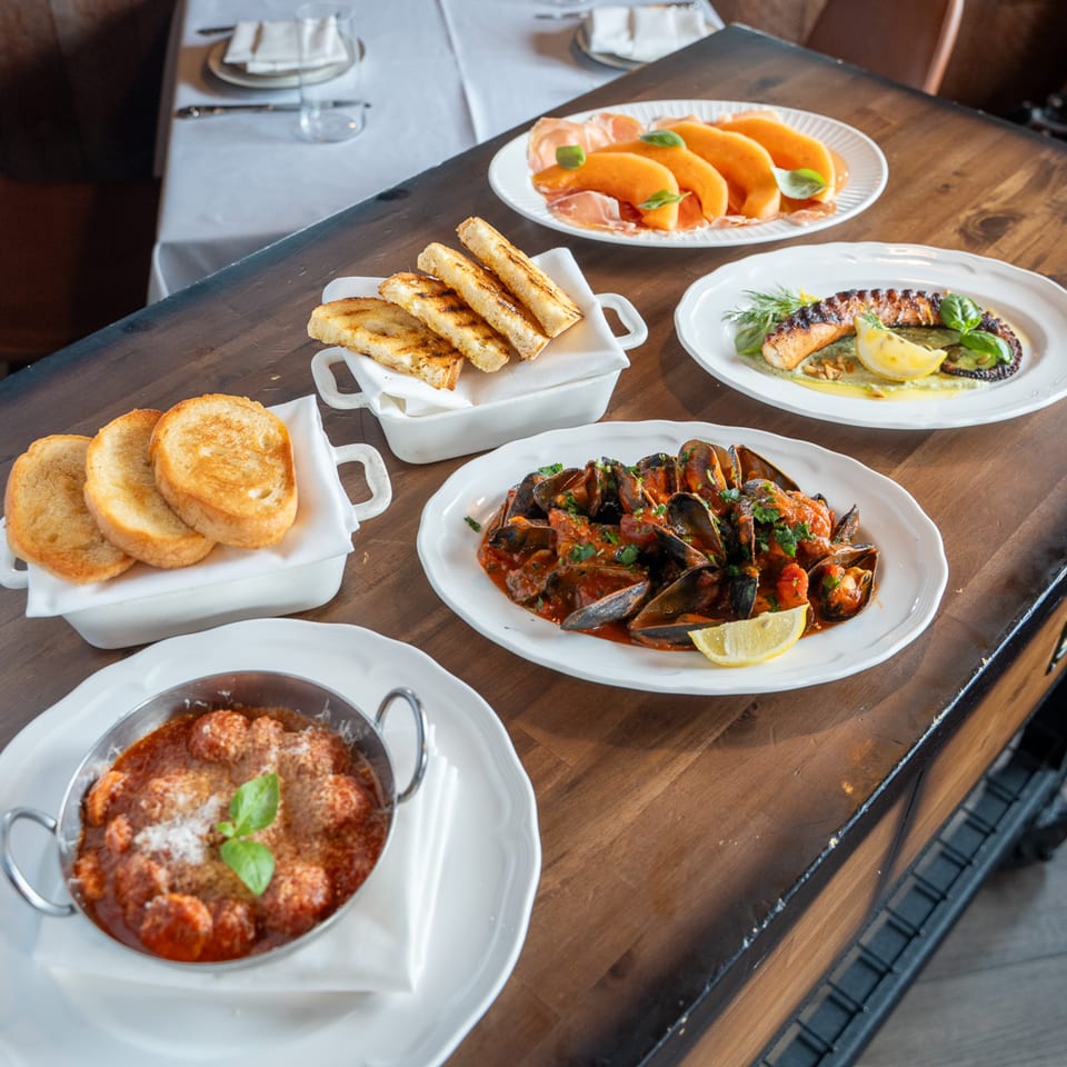 A wooden table laden with various Italian dishes, including mussels, meatballs, and grilled bread.
