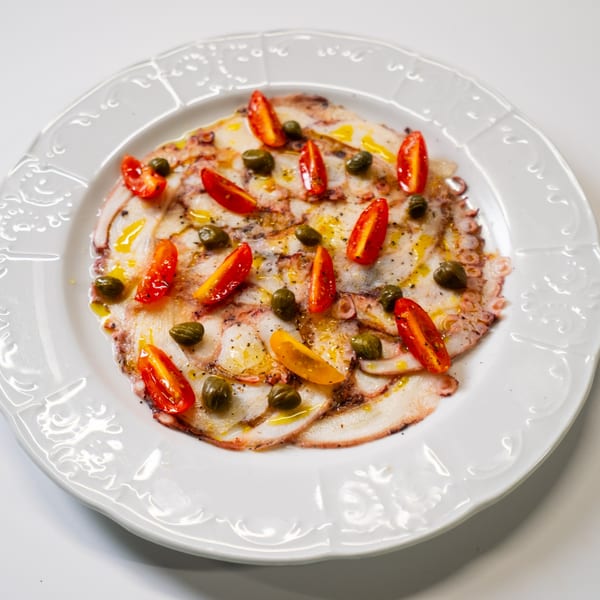 Octopus carpaccio with cherry tomatoes, capers, olive oil, and black pepper on a white plate.