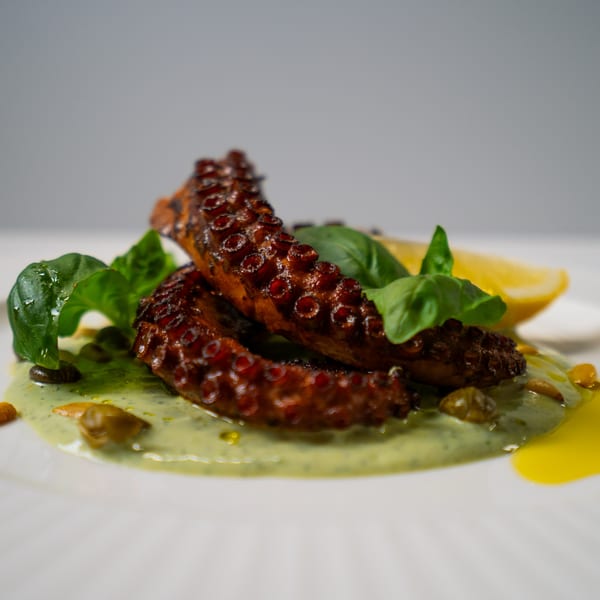 Grilled octopus tentacles on a bed of green sauce, garnished with basil and a lemon wedge.