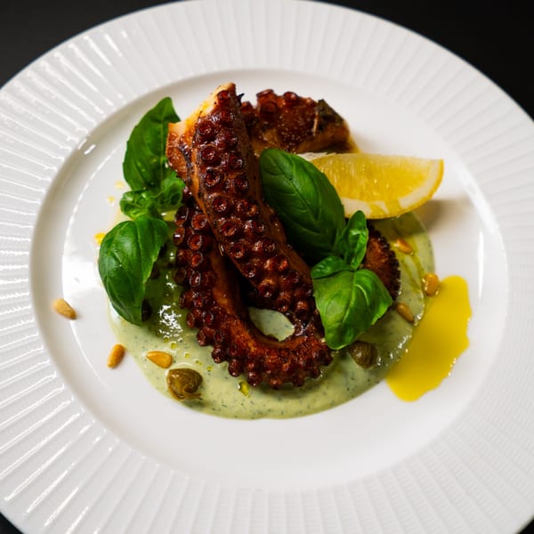 Grilled octopus with green sauce, fresh basil, pine nuts, capers, and a lemon wedge on a white plate.