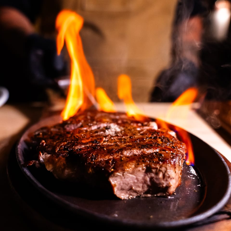 Flambéed steak on a sizzling cast iron plate, with bright orange flames rising from the dish.