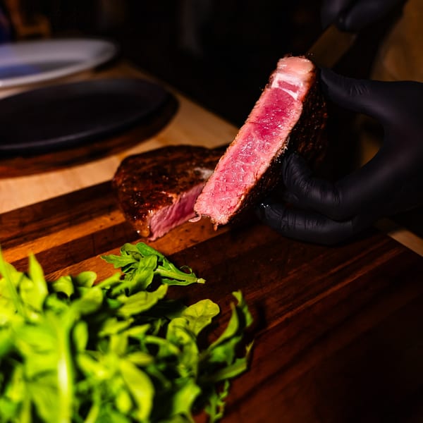 A gloved hand holds a perfectly cooked, sliced steak, revealing its pink interior, on a wooden board with greens.